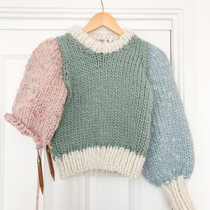 May include: A multi-colored knitted sweater with a green body, white trim, and pink and blue sleeves. The sweater is hanging on a wooden hanger.
