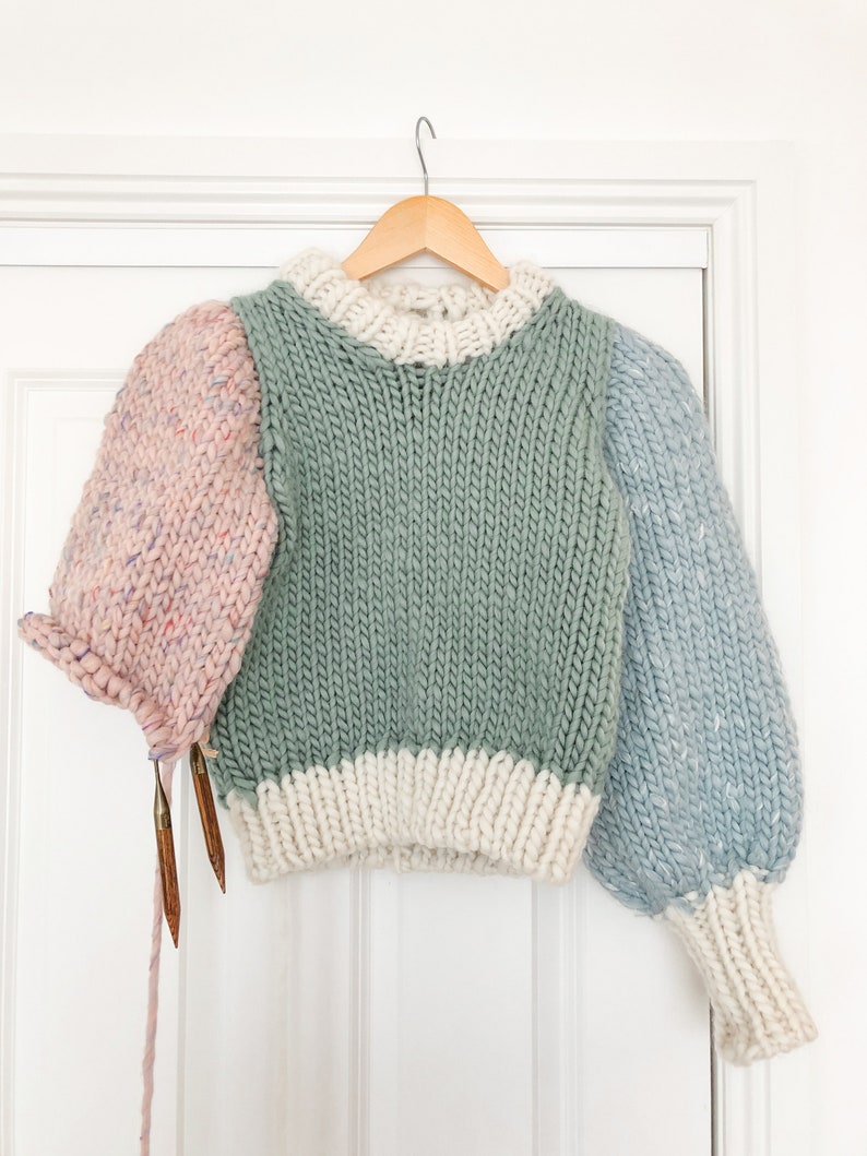 May include: A multi-colored knitted sweater with a green body, white trim, and pink and blue sleeves. The sweater is hanging on a wooden hanger.