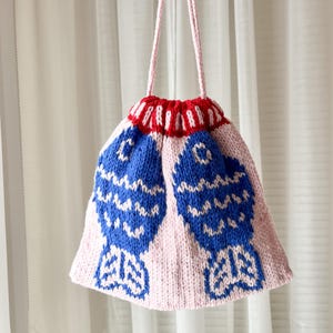 May include: A handmade knitted bag with a pink body and a red and white striped top. Two large blue fish designs are featured on the front. The bag has a pink drawstring closure and a pink strap.