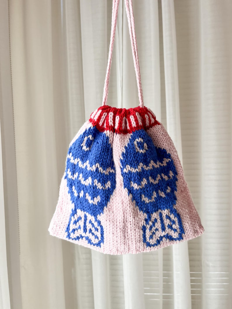 May include: A handmade knitted bag with a pink body and a red and white striped top. Two large blue fish designs are featured on the front. The bag has a pink drawstring closure and a pink strap.