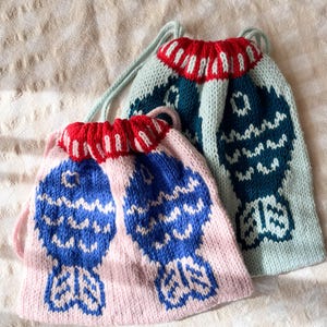 May include: Two knitted drawstring bags. One is light pink with blue fish designs, the other is teal with a similar design. Both have red and white striped trim at the top and light green drawstrings. The bags are on a beige surface.