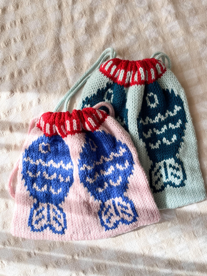 May include: Two knitted drawstring bags. One is light pink with blue fish designs, the other is teal with a similar design. Both have red and white striped trim at the top and light green drawstrings. The bags are on a beige surface.
