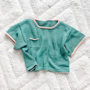 May include: A teal knitted short-sleeved shirt with a pink trim around the neckline and sleeves. The shirt has a small pocket on the front.
