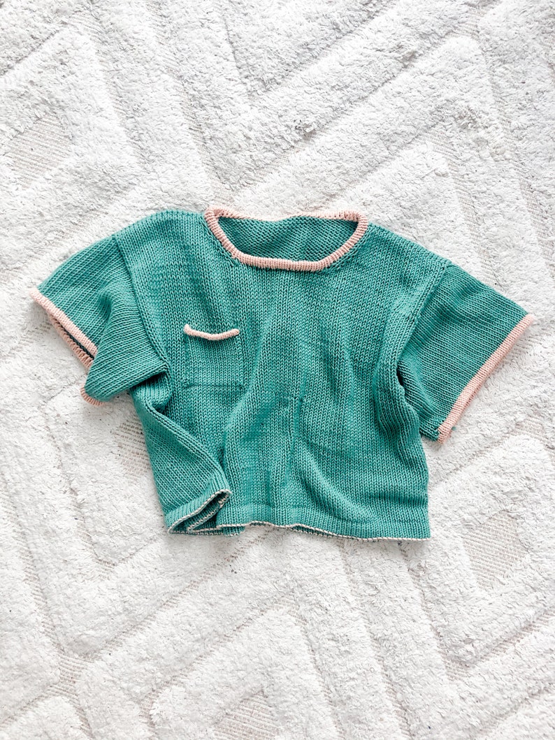 May include: A teal knitted short-sleeved shirt with a pink trim around the neckline and sleeves. The shirt has a small pocket on the front.