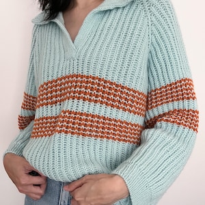 Könnte beinhalten: Hellblauer Strickpullover mit Kragen und horizontalem orange-weiß gestreiftem Design auf der Brust. Der Pullover hat lange Ärmel und eine gerippte Textur.