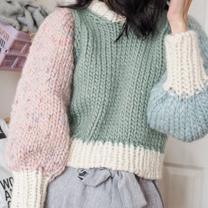 May include: A green, pink, and blue knitted sweater with a white trim. The sweater has a relaxed fit and a round neckline.
