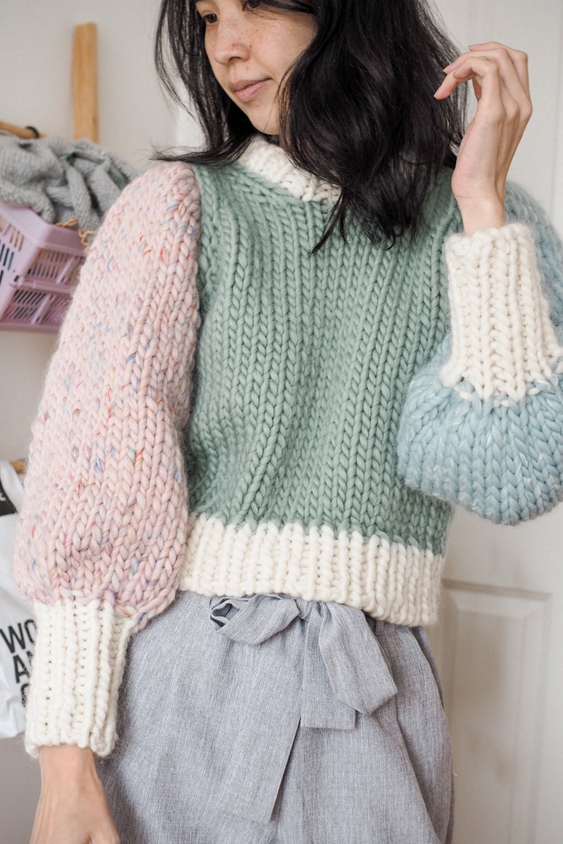 May include: A green, pink, and blue knitted sweater with a white trim. The sweater has a relaxed fit and a round neckline.