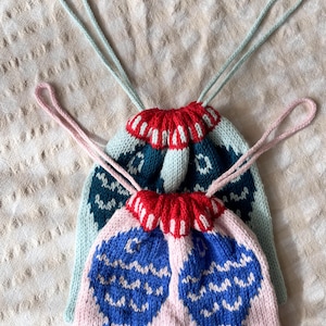 May include: Two knitted drawstring bags. One is light pink with blue and white fish-like designs, and the other is teal with a red and white scalloped top. Both have matching drawstring cords.