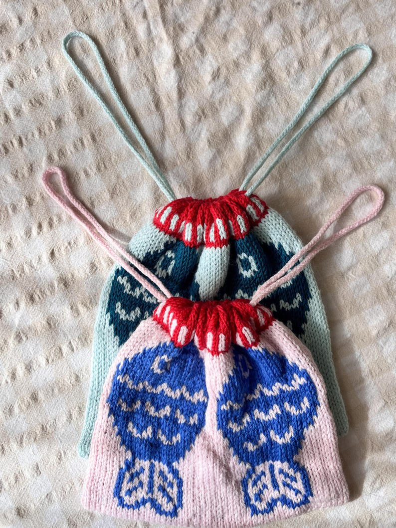 May include: Two knitted drawstring bags. One is light pink with blue and white fish-like designs, and the other is teal with a red and white scalloped top. Both have matching drawstring cords.