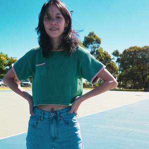 May include: A woman wearing a green cropped sweater with pink trim and blue jeans. She is standing on a blue and green tennis court.