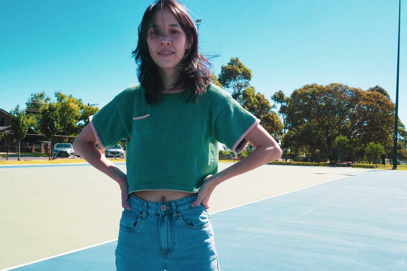 May include: A woman wearing a green cropped sweater with pink trim and blue jeans. She is standing on a blue and green tennis court.