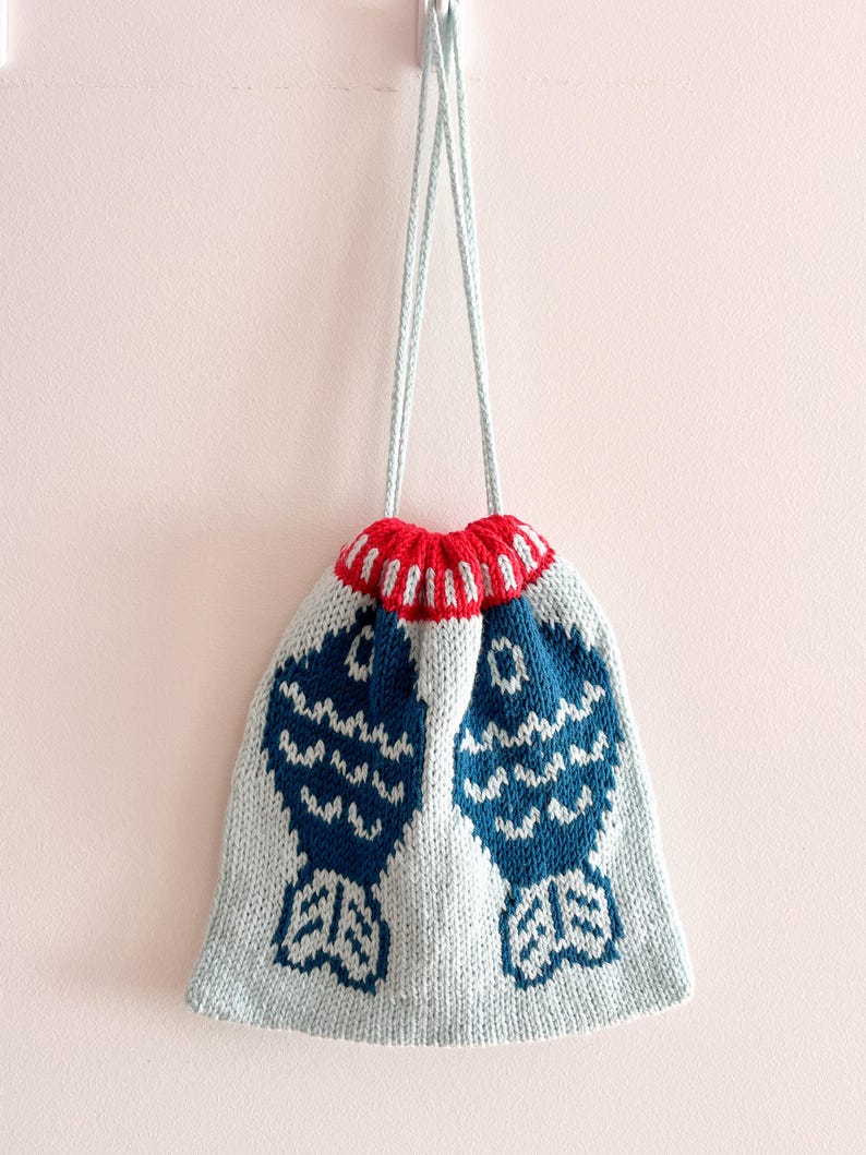 May include: A knitted drawstring bag with a light blue body and a red and white striped top. The bag features two large blue fish designs. The drawstring is light blue.