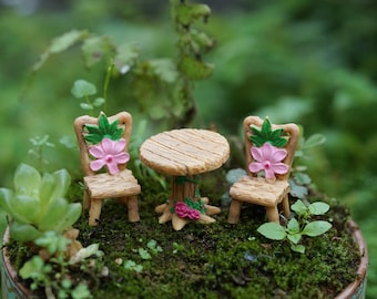 Può includere: Un tavolo e due sedie in legno in miniatura con fiori rosa e foglie verdi. Il set è perfetto per un giardino di fate o una scena in miniatura.