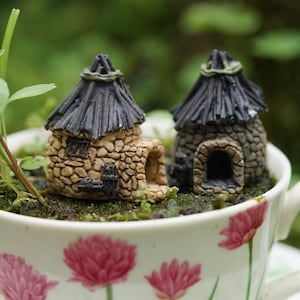 Op de afbeelding: Twee miniatuurstenen huizen met rieten daken, een met een ronde vorm en de andere met een meer rechthoekige vorm, zijn genesteld in een witte theekop met een bloemenpatroon.