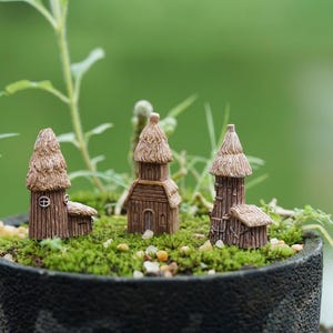 Peut inclure: Trois maisons miniatures en bois avec des toits de chaume, disposées dans un petit pot avec de la mousse verte et du gravier brun.