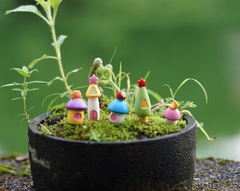 May include: A set of five miniature fairy houses made of ceramic. The houses are painted in various colors, including pink, yellow, blue, green, and white. They are decorated with small details, such as windows, doors, and roofs. The houses are arranged on a bed of green moss in a black pot.