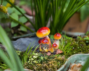May include: Three miniature ceramic mushrooms with red and yellow caps and white stems. The mushrooms are arranged in a cluster on a bed of green moss and grass.