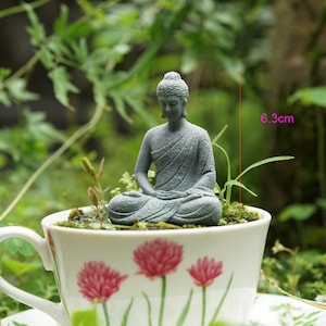 K&ouml;nnte beinhalten: Eine kleine graue Steinbuddha-Statue, die in einer wei&szlig;en Teetasse mit Blumenmuster sitzt. Die Teetasse ist 6,3 cm hoch.