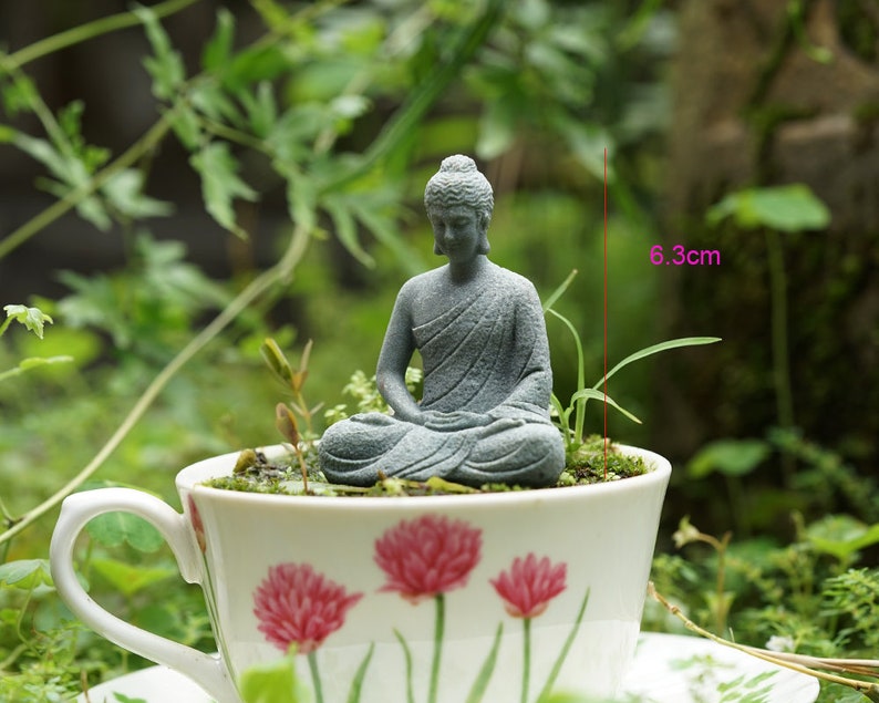 K&ouml;nnte beinhalten: Eine kleine graue Steinbuddha-Statue, die in einer wei&szlig;en Teetasse mit Blumenmuster sitzt. Die Teetasse ist 6,3 cm hoch.