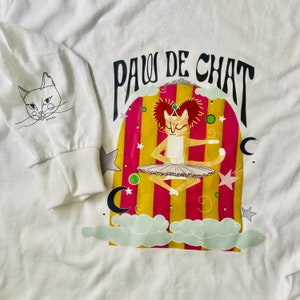 May include: White long-sleeve t-shirt with a colorful graphic of a cat wearing a tutu and the text "PAW DE CHAT".
