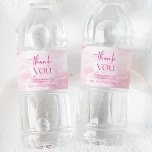 May include: Two clear plastic water bottles with pink and white labels that say "Thank You". The bottles are on a white lace doily.