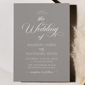 Op de afbeelding: Een grijze trouwuitnodigingskaart met witte kalligrafietekst, inclusief de namen Madison James en Nathaniel Dunn, en de datum. De kaart is geplaatst op een wit cirkelvormig oppervlak met een gedroogde plant op de achtergrond.