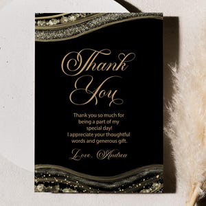 May include: A black thank you card with gold script and decorative gold and glitter accents. The card reads "Thank You" and includes a personalized message. The card is set against a neutral background with a decorative pampas grass.