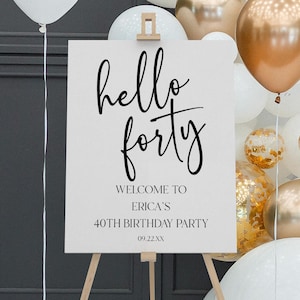 May include: A white sign on a wooden easel with the words "hello forty" in black script. The sign welcomes guests to Erica's 40th birthday party. Balloons in white and gold surround the sign, creating a festive atmosphere.