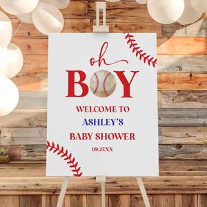 May include: A white sign on an easel with the words "oh BOY" in red, a baseball graphic, and "WELCOME TO ASHLEY'S BABY SHOWER 09.27.XX". Red baseball stitching accents the corners. White balloons and a wooden backdrop complete the scene.