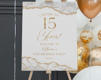 May include: A rectangular sign on a wooden tripod easel featuring a white background with gold marble-like edges. The text reads: Cheers to 15 Years! Welcome to Andrea's 15th Birthday Party. The sign is surrounded by white and metallic gold balloons.