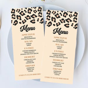 May include: Two beige menu cards with a leopard print design at the top, featuring a list of courses. The menus are on a white plate, with gold cutlery and a white napkin. A small vase of white flowers and a glass of water are also present.