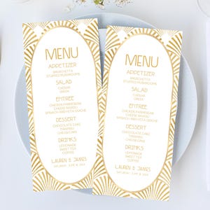 May include: Overhead shot of a table setting with two menu cards. The menus have a gold art deco design and the word "MENU" at the top. Gold utensils, a white napkin, and a small vase of flowers complete the elegant arrangement.