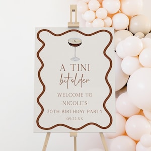 Może przedstawiać: Znak na drewnianym sztaludze z napisem "A Tini bit older" i "Welcome to Nicole's 30th Birthday Party". Znak ma brązową obwódkę i grafikę martini. Łuk balonowy znajduje się w tle.