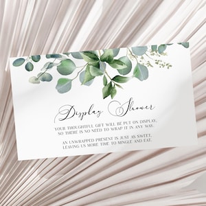 May include: A white card with green foliage and the text "Display Shower" in cursive. The text below reads "Your thoughtful gift will be put on display, so there is no need to wrap it in any way. An unwrapped present is just as sweet, leaving us more time to mingle and eat."
