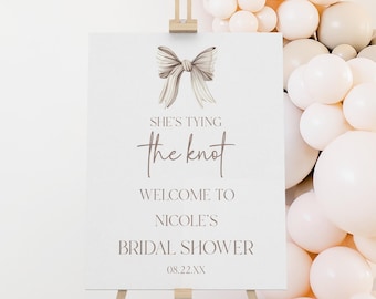 May include: A white sign with a brown bow and the text "She's tying the knot. Welcome to Nicole's Bridal Shower 08.22.XX" on a wooden easel in front of a wall decorated with a balloon arch.