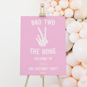 May include: A pink sign on a wooden easel with the text "BAD TWO THE BONE" and a skeleton hand making a peace sign. The sign welcomes guests to Chloe's 2nd birthday party.