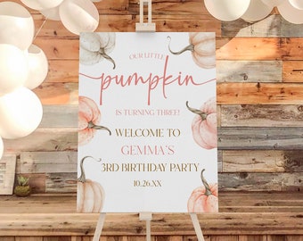 May include: A white easel displays a sign with the text "Our Little Pumpkin is Turning Three! Welcome to Gemma's 3rd Birthday Party." The sign features watercolor pumpkins in shades of peach and gray. White balloons and a wooden backdrop complete the scene.