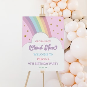 Może przedstawiać: Różowy i biały znak powitalny na drewnianym sztaludze. Na znaku widnieje napis "Olivia is on Cloud Nine" z tęczą i gwiazdami. Znak wita gości na 9. urodzinach Olivii.