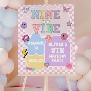 Op de afbeelding: Een kleurrijk verjaardagsfeestbord op een houten ezel. Het bord leest "NINE is a VIBE" met extra tekst: "Welcome to Olivia's 9th Birthday Party". Het bord heeft een geruite achtergrond, smiley's en andere decoratieve elementen.