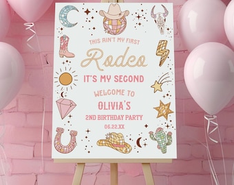 May include: A white rectangular birthday party sign on a wooden easel featuring the text "THIS AIN'T MY FIRST Rodeo IT'S MY SECOND WELCOME TO OLIVIA'S 2ND BIRTHDAY PARTY 06.22.XX" surrounded by illustrations of a cowboy hat, disco ball, boots, and cactus.