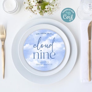 以下が含まれることがあります： 白いプレートに、青と白の雲のデザインが施され、「The bride is on cloud nine」と「Megan's Bridal Shower」の文字が書かれています。プレートは、白いテーブルの上に置かれ、金色のフォーク、ナイフ、スプーン、コップの水、小さな白い瓶が置かれています。