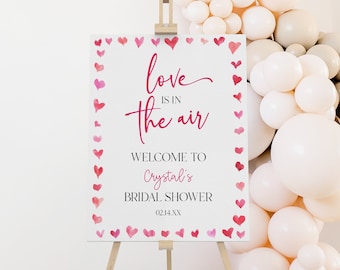 Könnte beinhalten: Ein weißes Schild mit einem rosa Aquarell-Herzrand und dem Text "love is in the air" in rosa Schrift. Das Schild trägt außerdem den Text "Welcome to Crystal's Bridal Shower 02.14.XX". Das Schild steht auf einem Holzstaffelei vor einer Wand aus weißen und beigefarbenen Luftballons.