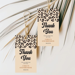 May include: Two rectangular gift tags with a tan background and a leopard print border. The tags read "Thank You" in a stylish font, with additional text below. The tags are tied with twine and are set against a white background with palm leaves.