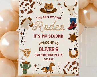 May include: A rectangular birthday party sign on a wooden easel featuring the text This Ain't My First Rodeo It's My Second Welcome To Oliver's 2nd Birthday Party 06.22.XX with illustrations of a cowboy hat, horse, cactus, and cow print.