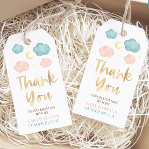 Op de afbeelding: Twee witte cadeaulabels met aquarel wolken en manen, met de tekst "Thank You" in gouden letters. De labels zitten in een kartonnen doos gevuld met versnipperd papier. De tekst op de labels luidt "For celebrating with us!" en "Kara & Glenn's Gender Reveal."