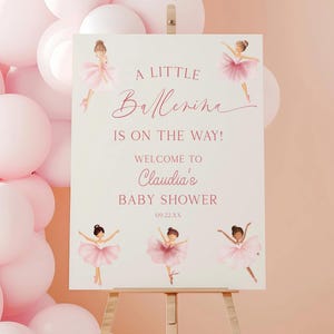 Könnte beinhalten: Ein Willkommensschild für eine Babyparty auf einer Staffelei mit dem Text "A Little Ballerina Is On The Way!" in rosa Schrift. Das Schild zeigt Illustrationen von Ballerinas in rosa Tutus und ist von rosa Ballons umgeben.