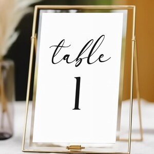 May include: A gold metal framed table number card with the word "table" in cursive and the number "1" in a bold font. The card is displayed on a white surface. A vase with dried flowers is in the background.