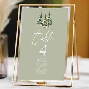 May include: A gold metal framed table number 4 sign. The sign has a light green background with a watercolor illustration of three pine trees. The word "table" is written in cursive, and the number 4 is in a bold font. Names are listed below.