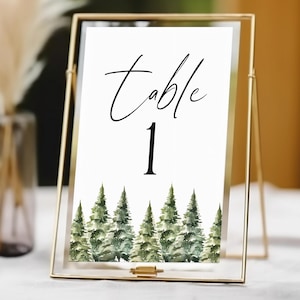 May include: A gold metal framed table number card. The white card has the word "Table" in elegant script above the number "1" in a bold font. The bottom of the card features a watercolor illustration of green pine trees. A vase with dried flowers is in the background.