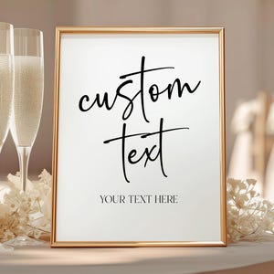 May include: A gold-framed sign with the words "custom text" in elegant script. Below, the text reads "YOUR TEXT HERE." Two champagne flutes and floral arrangements flank the sign, suggesting a celebratory event.